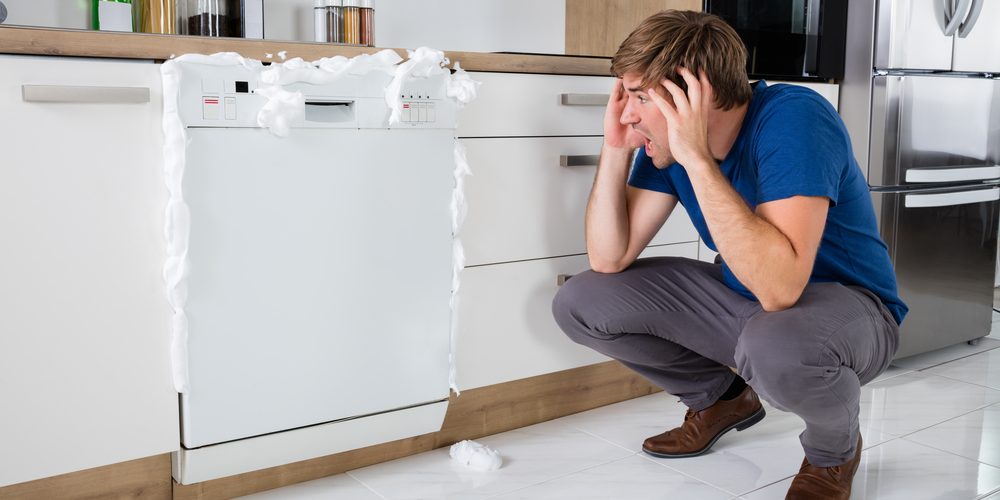 A man looks on in shock as soap emerges from the closed dishwasher in front of him