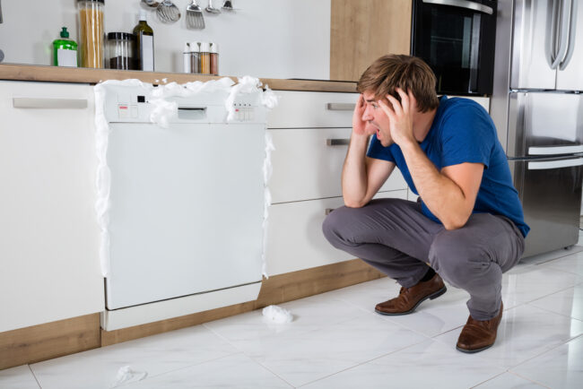 A man looks on in shock as soap emerges from the closed dishwasher in front of him
