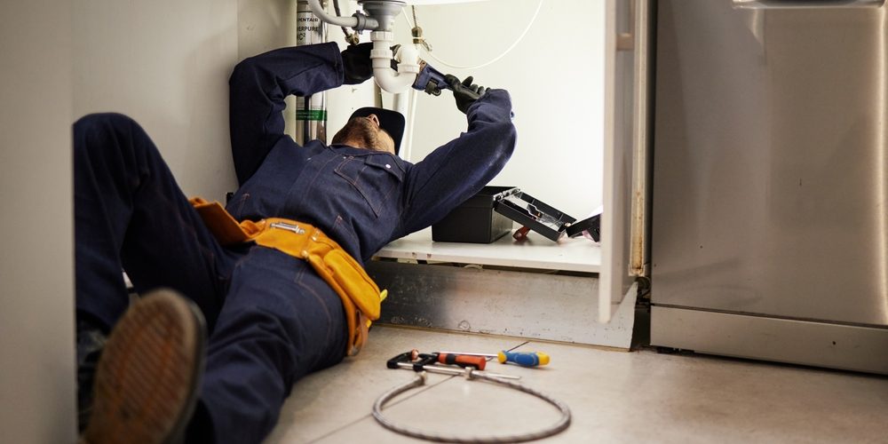 Kitchen,,Maintenance,And,Renovation,With,A,Plumber,On,The,Floor A plumber uses a wrench to fix a pipe under a kitchen sink